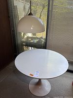 Photo showing white round tulip table under hanging pendant light near window blinds.