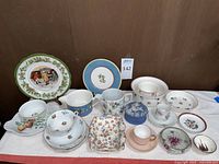 Wide view of multiple porcelain and ceramic items on white surface, including cups, saucers, decorative plates and bowls with floral, bird and countryside themes.