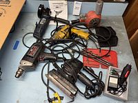 Six used power tools including a blue Chicago Electric angle grinder, a yellow Craftsman drill, an electric sander, and a few other power tools with cords and bits on a blue surface