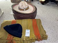 Lot showing all three items: sombrero placed on dark wood box, vintage serape folded with fringes visible, and dark blue wool beanie resting on serape.