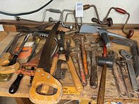 Overview of vintage hand tools on wooden surface including saws, brace drills, wrenches, hammers, tin snips, and pliers. Many tools show rust and use wear.