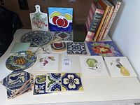Photo showing a variety of ceramic tiles with colorful designs of fruits, flowers, and birds, alongside a collection of cookbooks and a tin tray.