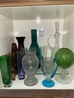 Photo showing all 11 glass pieces arranged inside a cabinet shelf, various shapes and colors visible.