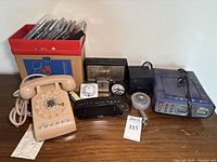 Photo of vintage electronics and containers showing rotary phone, various clocks, and Taylor weather station with CDs in a red and brown box.
