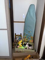 Photo of the ironing board standing upright behind a box containing several vintage plush toys and a small hand-painted chair next to the ironing board.