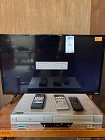 Full set showing the 32-inch Vizio flat screen TV positioned behind and Zenith dual DVD/VHS player below with the three remotes in front. TV displays a label and screen is off.