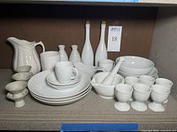 Photo showing full set of white ceramic serving dishes including plates, cups, pitchers, egg cups, mortar and pestle, and cruet bottles arranged on shelf.