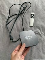 Apple TV box held in hand with power cable and white remote control visible on a fabric surface