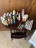 Wide view of assorted acrylic, craft paints, Mod Podge bottles, brushes, and spray paint can arranged on a wooden table.