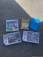 Photo showing the Sorry game box, Chinese Checkers box, two 1000-piece puzzles (Music and Movie themes), and blue puzzle mat laid out on ground.