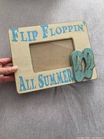Front view of the rectangular wooden picture frame held by hand, showing the cream color with turquoise blue lettering 'FLIP FLOPPIN' ALL SUMMER' and a turquoise wooden flip-flop decoration on the right corner.