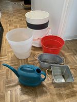 Photo showing five assorted plant pots including white plastic pots with and without black band, a red pot, and two metal planters, along with a blue plastic watering can on a parquet floor.