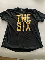 Front view of black T-shirt with gold 'THE SIX' print. Shows zipper on lower left side.