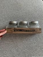 Set of three new jam jars with lids in cardboard and plastic packaging, showing front and sides of jars.