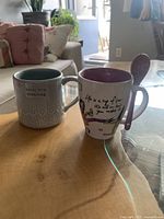 Two ceramic mugs on wooden table, one with embossed heart details and message, other taller mug with attached spoon and text