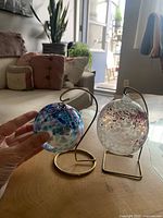 Two hanging glass spheres on metal stands displayed on a wooden surface with a background showing a living area and outside light.