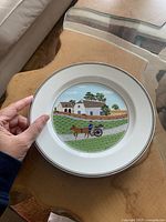 Porcelain decorative plate showing country scene with carriage and buildings, held by hand for scale on wooden table