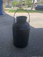 Front view of the black metal milk jug showing its shape, handle, and surface dents.