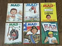 Six vintage Mad magazines arranged in two rows. Covers show Alfred E. Neuman in various humorous scenes, including brushing teeth, sports, and military themes. Condition shows slight age wear.