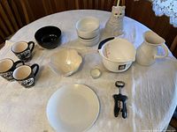 Overview of entire ceramic set including mugs, bowls, pitcher, cat figurine, plate, dish, candle holder, and a corkscrew bottle opener on a white tablecloth.