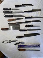 Varied cutting utensils laid out including knives with plastic and wooden handles, cleaver, kitchen shears, and sharpeners on round tablecloth.