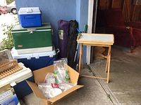 Camping items arranged including coolers stacked, camping chairs in carry bags, folding bar table and box with disposable cups and utensils in cardboard box.