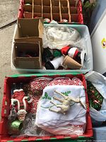 Overview of multiple red and green plastic crates containing various vintage Christmas ornaments, holiday linens, and decorations such as Santa figures and a ceramic manger scene.