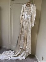 Full length view of ivory satin wedding dress hanging on door, showing long train and structured shoulders and sleeves.