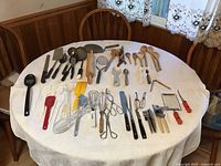 Table full of assorted kitchen utensils arranged on a white tablecloth showcasing wooden, metal, and plastic cooking tools mostly vintage and lightly used.