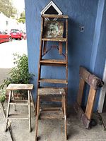 Wooden ladders and wooden dolly/cart displayed against blue wall outside with some background cars