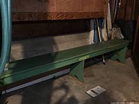 Long green garden bench with sturdy triangular legs, placed indoors on a concrete floor.