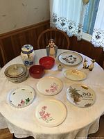 View of the ceramic and porcelain items arranged on a table with lace tablecloth, showing jars, plates with floral designs, figurine vase, and ceramic bowls.