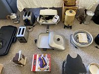 Group of vintage and modern kitchen appliances including toasters, griddles, coffee makers, electric grills, and kettles arranged on a carpeted floor.