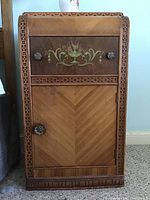 Front view of one bedside table showing carved trim, parquetry chevron veneer, painted motif and knob pulls