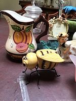 Photo showing ceramic pitcher with fruit design and a bee figurine with metal legs