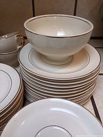 Stacked vintage white plates with thin gold rims. Large matching pedestal bowl and teacups with gold trims visible.