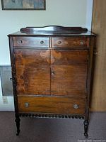 Front view of vintage tall wooden dresser showing dark wood with lighter wood detailing and decorative hardware.