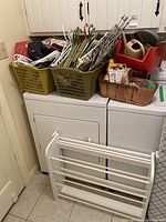 View of assortment of household items including multiple clothes hangers in green and white plastic baskets, plus an orange basket with various items on top of a washer and dryer.