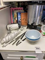 Photo showing coffee urn, electric hand mixer, three mixing bowls, cookbooks, and metal nut chopper on white appliance surface