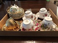Multiple bone china cups and saucers displayed in a box, showing a variety of floral patterns and gold-trimmed edges.