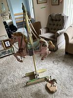 Large pink carousel horse mounted on a cream wooden stand with ribbons, standing on carpeted floor in a living room setting with chairs and framed pictures.