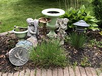 Garden set displayed outdoors in a flower bed showing all items together including statues, planters, sundial, and stepping stones.
