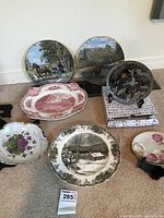 Six assorted decorative plates and dishes arranged on floor, including horse and carriage collector plates, black bird design plate, winter cabin scene plate, and two floral serving dishes.