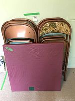Photo of four folded pink metal frame chairs and one with patterned back and folding table with dark red padded top.