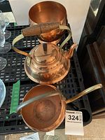 Copper planter, kettle with wooden handle, and watering can with arched brass handle displayed on a shelf with other household items.