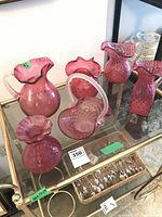 Six pieces of cranberry glass displayed on a glass cabinet including a brides basket with applied handle and five lemonade pitchers with diamond patterns.