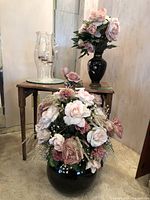 Two artificial flower arrangements displayed on and beside a small wooden table. One taller arrangement in a black vase and a larger round arrangement in a black vase. Clear glass hurricane lamp with three candle holders sitting on the table.