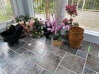 Overview of the lot showing multiple bunches of faux flowers, black metal scuttle vase, and wicker containers on tiled floor near window
