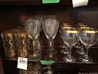Shelf display of assorted glasses, including tall gold filigree patterned glasses, stemmed wine glasses with gold rings on stems, and goblets with gold patterned rims.