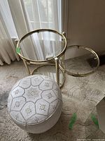 Glass top table with gold-tone metal frame and round surfaces; white embroidered pouf beside it.